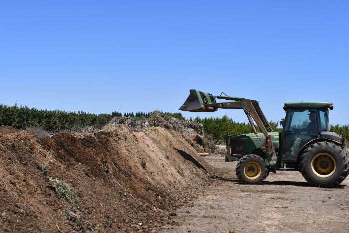 An image of a large brown compost pile next to a tractor with a loader, lifting up compost into the air.
