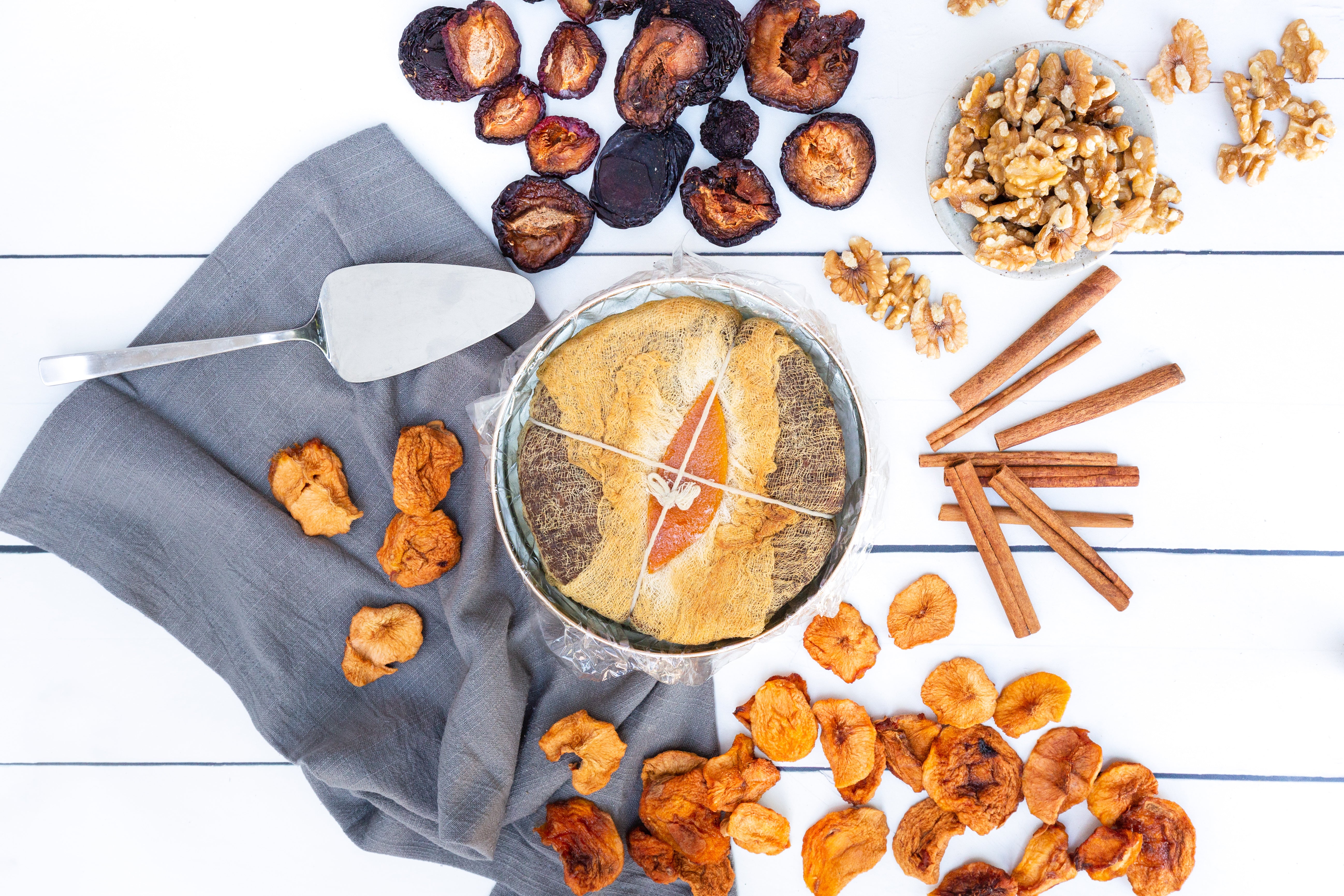 A flatlay of a fruitcake wrapped in cheescloth, surrounded by dried fruit slices, walnuts, and cinnamon sticks.
