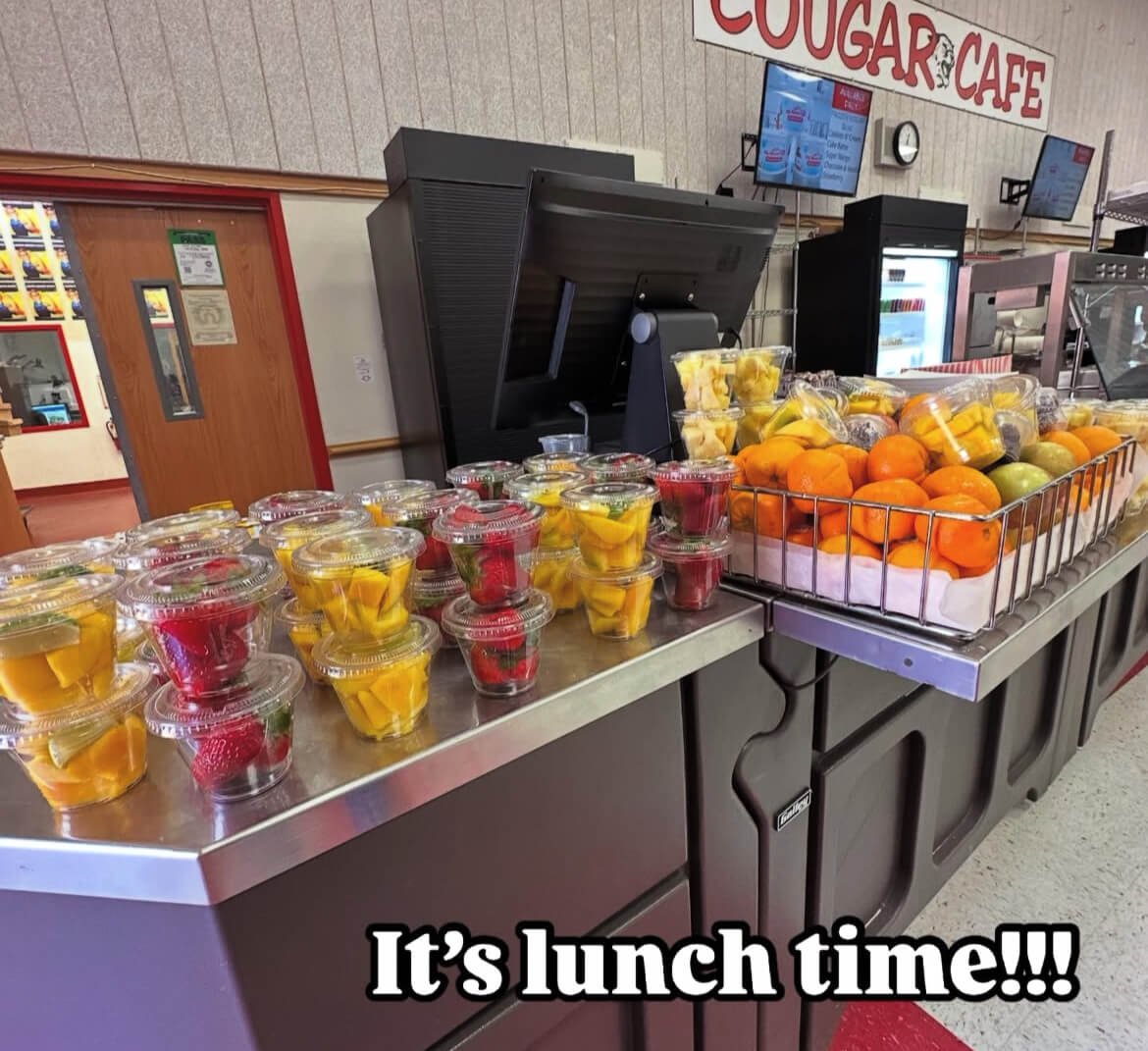 An image of a school cafeteria, and a table covered with colorful packaged fruit, including mango, straberries, pineapple, oranges, and more.