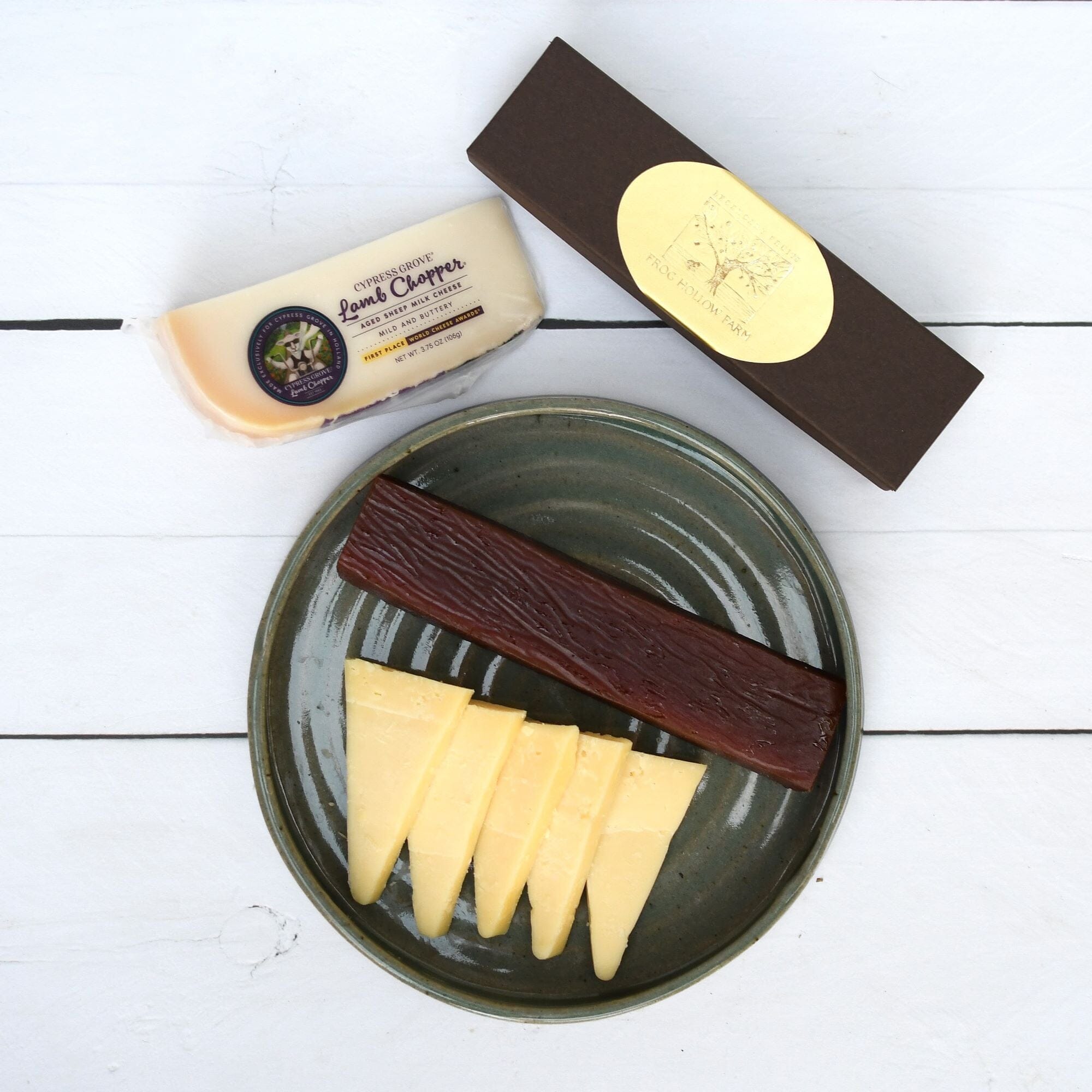 A flatlay of a block of Lamb Chopper cheese, a Frog Hollow gift box, and a plate filled with cheese slices and quince paste.