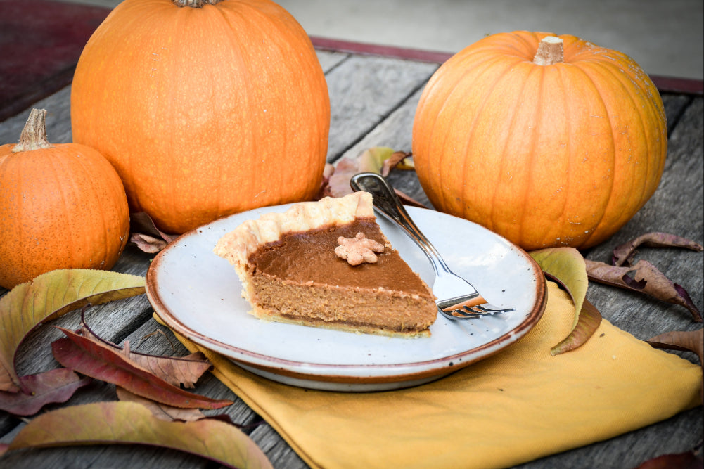 A slice of golden brown pumpkin pie, sitting on a plate, surrounded by decorative pumpkins and fall leaves.